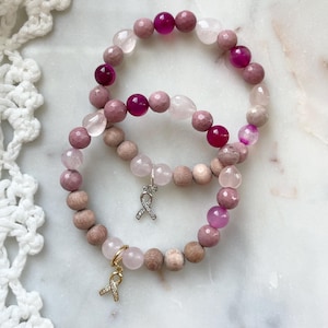 Puede incluir: Dos pulseras de cuentas con cuentas rosas, moradas y de madera. Cada pulsera presenta un dije en forma de lazo. Las pulseras se exhiben sobre una superficie de mármol blanco.