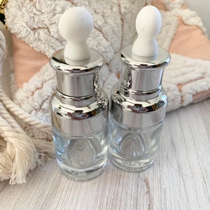 Puede incluir: Dos botellas de vidrio transparente con tapas plateadas y goteros blancos. Las botellas est&aacute;n sobre una superficie de madera clara.