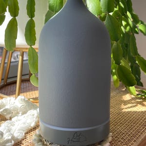 Puede incluir: Un difusor de aceites esenciales gris con una parte superior redondeada y una base de color claro. El difusor est&aacute; sobre una superficie tejida y rodeado de plantas verdes. El texto "Oil Haven" est&aacute; impreso en la base.