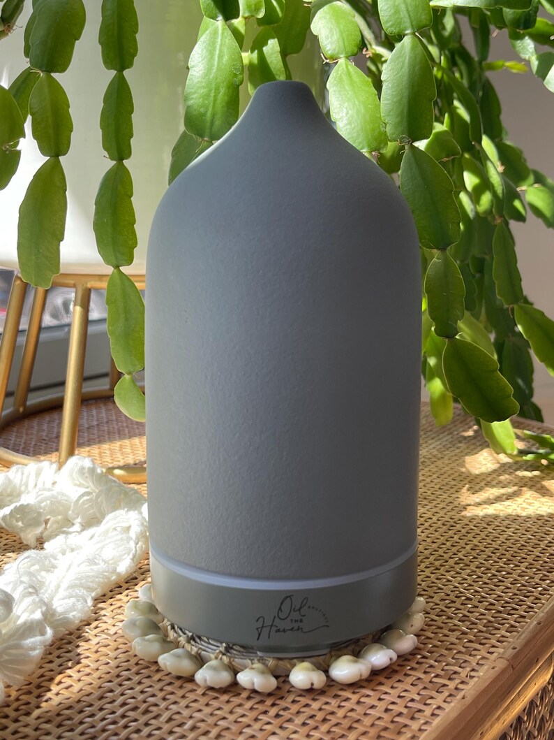 Puede incluir: Un difusor de aceites esenciales gris con una parte superior redondeada y una base de color claro. El difusor est&aacute; sobre una superficie tejida y rodeado de plantas verdes. El texto "Oil Haven" est&aacute; impreso en la base.