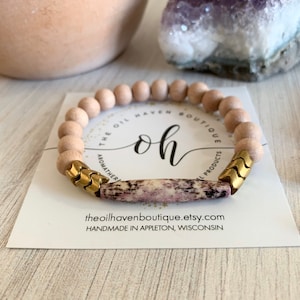 Op de afbeelding: Een houten kralen armband met een gouden metalen bedel en een roze en witte steen accent. De armband ligt op een witte kaart met de tekst "The Oil Haven Boutique" en "Handmade in Appleton, Wisconsin".