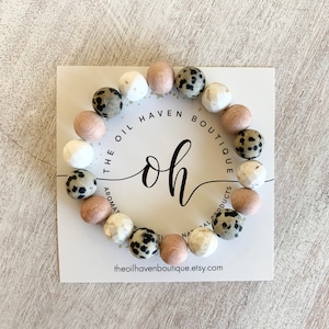 Puede incluir: Un brazalete de silicona con cuentas marrones, blancas y negras. El brazalete tiene un logotipo de "The Oil Haven Boutique" con el texto "oh" en el centro.