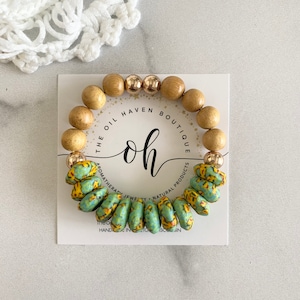 May include: A wooden bracelet with gold beads and a turquoise and yellow patterned bead. The bracelet is on a white card with the text "The Oil Haven Boutique oh Aromatherapy Jewelry Natural Products The Oil Haven Boutique"