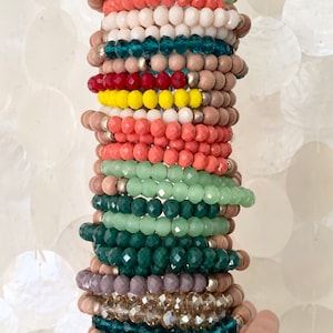 May include: A stack of colourful beaded bracelets in shades of green, yellow, red, pink, and brown. The bracelets are made of glass and wood beads.