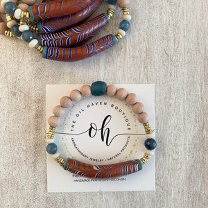 May include: A wooden bracelet with blue and white beads, a brown and blue patterned ceramic bead, and gold accents. The bracelet is on a white card with the text "THE OIL HAVEN BOUTIQUE oh AROMATHERAPY JEWELRY NATURAL PRODUCTS the handmade in america, wisconsin".