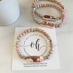 Pode incluir: Duas pulseiras feitas com contas de madeira natural e detalhes de concha. As pulseiras estão empilhadas uma em cima da outra e são exibidas em um cartão branco com o texto "The Oil Haven Boutique, Aromatherapy & Natural Products, theoilhavenboutique.etsy.com, Handmade in Appleton, Wisconsin".