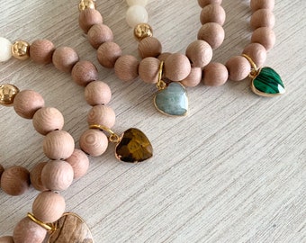 May include: Three wooden bead bracelets with heart-shaped gemstone charms. The bracelets are layered on a white wooden surface. The charms are green, brown, and light blue.
