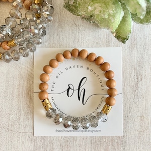 Könnte beinhalten: Ein Armband aus Holzperlen mit klaren Kristallperlen und goldenen Akzenten. Das Armband trägt den Text "The Oil Haven Boutique" und "Oh". Der Text "Aromatherapy Jewelry - Natural Products" befindet sich ebenfalls auf dem Armband. Das Armband liegt auf einer weißen Karte mit der Website "theoilhavenboutique.etsy.com".