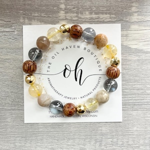 Può includere: Un braccialetto di perline con una varietà di perline di pietra naturale nei toni del marrone, dell'oro e del trasparente. Il braccialetto è esposto su una carta bianca con il testo "The Oil Haven Boutique" e "Handmade in Appleton, Wisconsin."