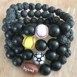 May include: Stack of beaded bracelets featuring sports-themed beads. The bracelets include black matte beads, lava rock beads, and beads shaped like a baseball, softball, soccer ball, and football.