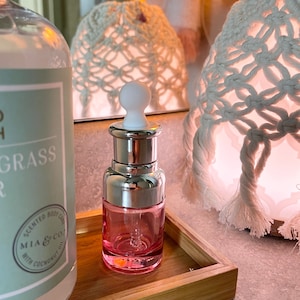Puede incluir: Una botella de vidrio rosa con tapa plateada y gotero. La botella est&aacute; sentada en una bandeja de madera. La botella est&aacute; etiquetada con el texto "MIA & CO. SCENTED BODY OIL WITH COCONUT OIL".