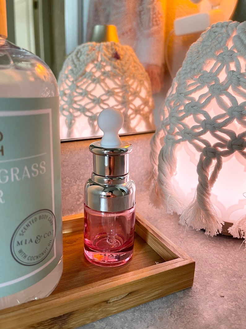 Puede incluir: Una botella de vidrio rosa con tapa plateada y gotero. La botella est&aacute; sentada en una bandeja de madera. La botella est&aacute; etiquetada con el texto "MIA & CO. SCENTED BODY OIL WITH COCONUT OIL".