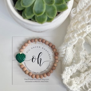 Puede incluir: Un brazalete de cuentas de madera con un dije de corazón verde. El brazalete está hecho por Oil Haven Boutique y presenta el texto "Oil Haven Boutique oh Aromatherapy Jewelry Natural Products theoilhavenboutique.etsy.com Handmade in Appleton, Wisconsin".