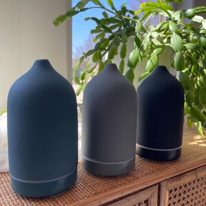 Puede incluir: Tres difusores de aceites esenciales en tonos azul, gris y negro se exhiben sobre un mueble tejido. Cada difusor tiene una parte superior redondeada y una base con una luz sutil. Los difusores est&aacute;n etiquetados con el texto "Haver".