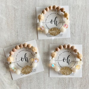 Puede incluir: Tres pulseras de madera con cuentas blancas y beige, detalles dorados y piedras iridiscentes. Las pulseras están apiladas una encima de la otra y presentan el texto "The Oil Haven Boutique" y "oh".