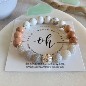 Może przedstawiać: Bransoletka z koralików z białymi, jasnobrązowymi i szarymi koralikami. Bransoletka zawiera srebrne akcenty i jest prezentowana na białej karcie z napisem "The Oil Haven Boutique".