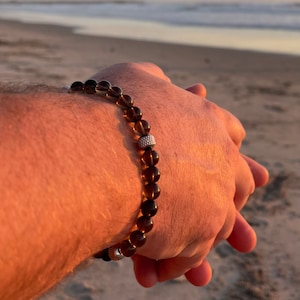 May include: A brown beaded bracelet with a silver accent on a person's wrist. The bracelet is on a beach with sand and ocean in the background.