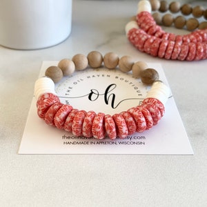 May include: A red and white patterned bracelet with wooden beads. The bracelet is handmade and has the text "Oh" and "The Oil Haven Boutique" on the card it is displayed on.
