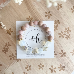 May include: A bracelet made with natural wood and white beads. The bracelet has a gold charm with the text "The Oil Haven Boutique" and "Aromatherapy Jewelry - Natural Products". The bracelet is handmade in Appleton, Wisconsin.