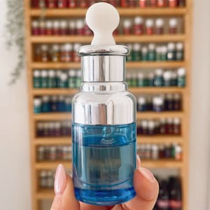 Puede incluir: Un peque&ntilde;o frasco de vidrio transparente con una tapa plateada y un gotero blanco. El frasco est&aacute; lleno de un l&iacute;quido azul claro. El frasco est&aacute; siendo sostenido en una mano.