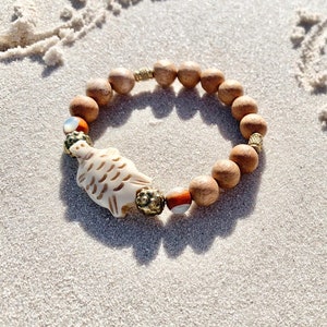 Könnte beinhalten: Ein Armband aus Holzperlen mit einem geschnitzten Fisch-Anhänger und goldenen Akzenten. Das Armband liegt auf einer weißen Sandfläche.