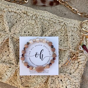 Puede incluir: Una pulsera de cuentas de madera, perlas y piedras preciosas descansa sobre un bolso de paja tejido. La pulsera va acompañada de una tarjeta que dice "The Oil Haven Boutique" y "Handmade in Appleton, Wisconsin."
