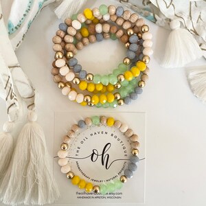 Pode incluir: Um conjunto de cinco pulseiras com contas de madeira, ouro e vidro em tons de amarelo, verde e cinza. As pulseiras estão empilhadas umas sobre as outras e são exibidas em um fundo branco com um cartão de visita que diz "The Oil Haven Boutique, Joias de aromaterapia, Produtos naturais, theoilhavenbou...e.etsy.com, Feito à mão em Appleton, Wisconsin".