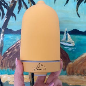 Puede incluir: Un difusor de aceites esenciales amarillo y beige con una parte superior redondeada y una base. El texto "Oil... Haven" est&aacute; impreso en la base. El fondo presenta una pintura de una escena tropical con palmeras y un velero.