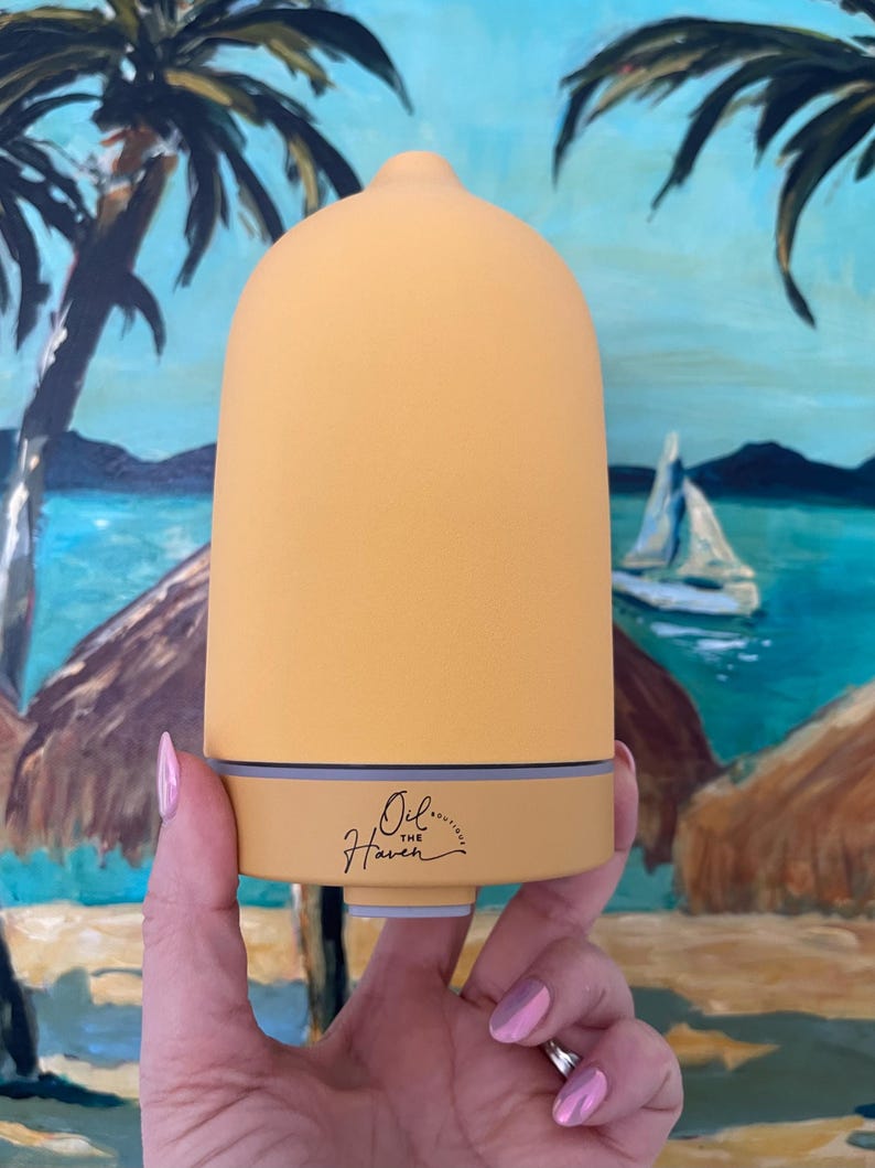 Puede incluir: Un difusor de aceites esenciales amarillo y beige con una parte superior redondeada y una base. El texto "Oil... Haven" est&aacute; impreso en la base. El fondo presenta una pintura de una escena tropical con palmeras y un velero.