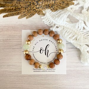 Könnte beinhalten: Ein Armband aus Holzperlen mit goldenen Perlen und grünen Edelstein-Akzenten. Das Armband befindet sich auf einer weißen Karte mit dem Text "THE OIL HAVEN BOUTIQUE oh AROMATHERAPY JEWELRY NATURAL PRODUCTS theoilhavenue.etsy.com HANDMADE IN APPLETON, WISCONSIN".