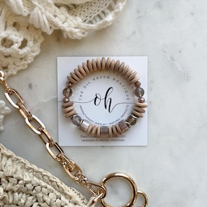 May include: A wooden bead bracelet with clear and brown beads, displayed on a white card with the text "THE OIL HAVEN BOUTIQUE". A gold chain and a straw bag are in the foreground. A red chili pepper charm hangs from the chain.