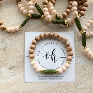 Pode incluir: Um conjunto de três pulseiras com contas de madeira e vidro. As pulseiras são de cor marrom, bege e branco com detalhes verdes. As pulseiras estão empilhadas sobre uma superfície de madeira. O cartão no centro diz "The Oil Haven Boutique, oh, Aromatherapy Jewelry, Natural Products, theoilhavenboutique.etsy.com, Handmade in Appleton, Wisconsin."