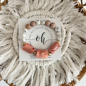 Könnte beinhalten: Ein rosa-weißes Perlenarmband mit dem Text "The Oil Haven Boutique" und "oh" in einem goldenen Kreis. Das Armband ist handgefertigt aus natürlichen Produkten.