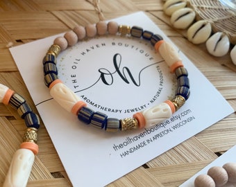Può includere: Primo piano di un braccialetto realizzato con legno naturale, perline a strisce blu e bianche e perline in osso intagliate con accenti pesca. Il braccialetto è su una carta bianca con il testo "The Oil Haven Boutique, Aromatherapy Jewelry, Natural Products, theoilhavenboutique.etsy.com, Handmade in Appleton, Wisconsin".