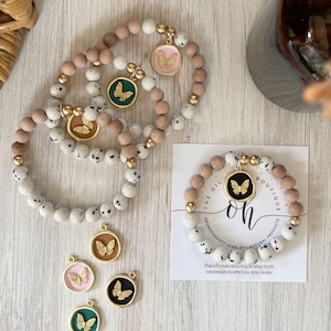 Puede incluir: Tres pulseras blancas y marrones moteadas con dijes de mariposa dorados. Las pulseras están sobre una superficie de madera con una tarjeta que dice "The Oil Haven Boutique" y tiene un dije de mariposa dorado con la palabra "oh" en ella.