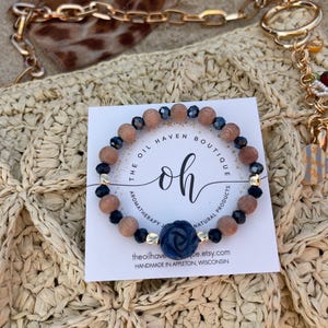 May include: A beaded bracelet with wooden and dark blue beads, accented with gold-colored spacers and a dark blue rose. The bracelet is displayed on a woven surface with a tag that reads "The Oil Haven Boutique".