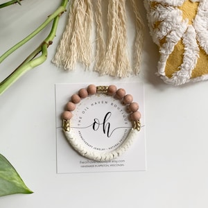May include: A white and gold beaded bracelet with the text "The Oil Haven Boutique" and "Oh" on the card. The bracelet is made with natural products and aromatherapy jewelry.