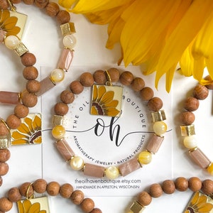 May include: Three wooden bead bracelets with sunflower charms. Each bracelet features a different color of beads, including brown, beige, and gold. The bracelets are displayed on a white surface with a business card that reads "The Oil Haven Boutique, aromatherapy jewelry, natural products, theoilhavenboutique.etsy.com, handmade in Appleton, Wisconsin."
