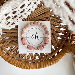 Puede incluir: Un brazalete de cuentas de madera con un dije de metal dorado y una banda con estampado rosa y blanco. El brazalete está sobre una tarjeta blanca con el texto "The Oil Haven Boutique", "oh", "Aromatherapy Jewelry - Natural Products", "theoilhaven.com", y "Handmade in Appleton, Wisconsin".