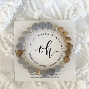 May include: Gray and brown beaded bracelet with a gold heart charm. The bracelet is on a white card with the text "The Oil Haven Boutique oh Aromatherapy Jewelry Natural Products theoilhavenboutique.com Handmade in Appleton, Wisconsin".