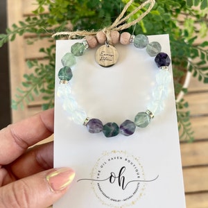 May include: A green, purple, and white crystal bracelet with a silver charm that says "Exercise Patience". The bracelet is on a white card with the text "The Oil Haven Boutique" and "Handmade in Appleton, Wisconsin".