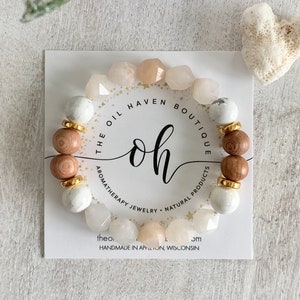 Pode incluir: Um bracelete de contas brancas, cor-de-rosa e castanhas com espaçadores dourados. O bracelete está sobre um cartão branco com o texto "THE OIL HAVEN BOUTIQUE oh AROMATHERAPY JEWELRY NATURAL PRODUCTS theo. 3M HANDMADE IN APPLETON, WISCONSIN".