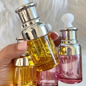 Puede incluir: Cuatro botellas de vidrio con gotero, con tapas plateadas y goteros blancos. Dos botellas son amarillas, una es rosa y una es transparente.