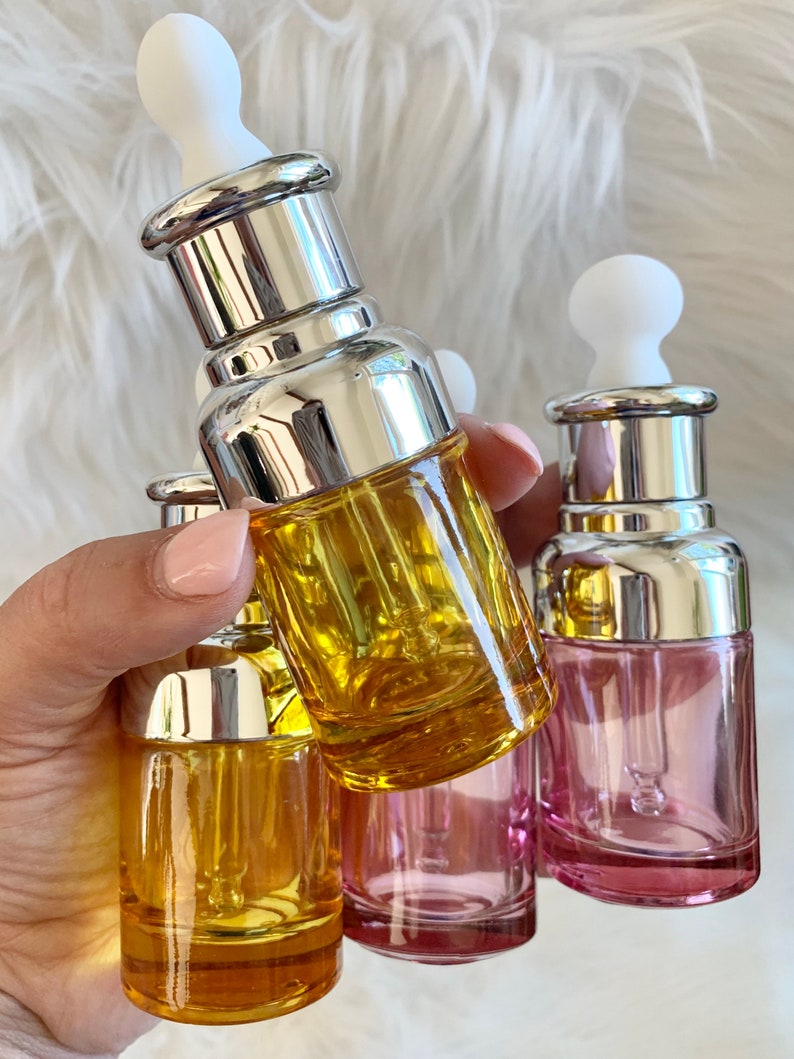 Puede incluir: Cuatro botellas de vidrio con gotero, con tapas plateadas y goteros blancos. Dos botellas son amarillas, una es rosa y una es transparente.