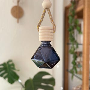 May include: A dark blue, diamond-shaped glass bottle hanging from a braided tan rope with gold metallic threading. The bottle features a light-colored, ribbed wooden cap and a small, round wooden bead accent on the suspension cord.