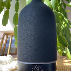 Puede incluir: Un difusor de aceites esenciales gris oscuro con una superficie texturizada y una parte superior redondeada. La base es negra con el texto "Oil the Haven" en escritura dorada. El difusor se encuentra sobre una superficie de cuentas blancas, con plantas verdes en el fondo.