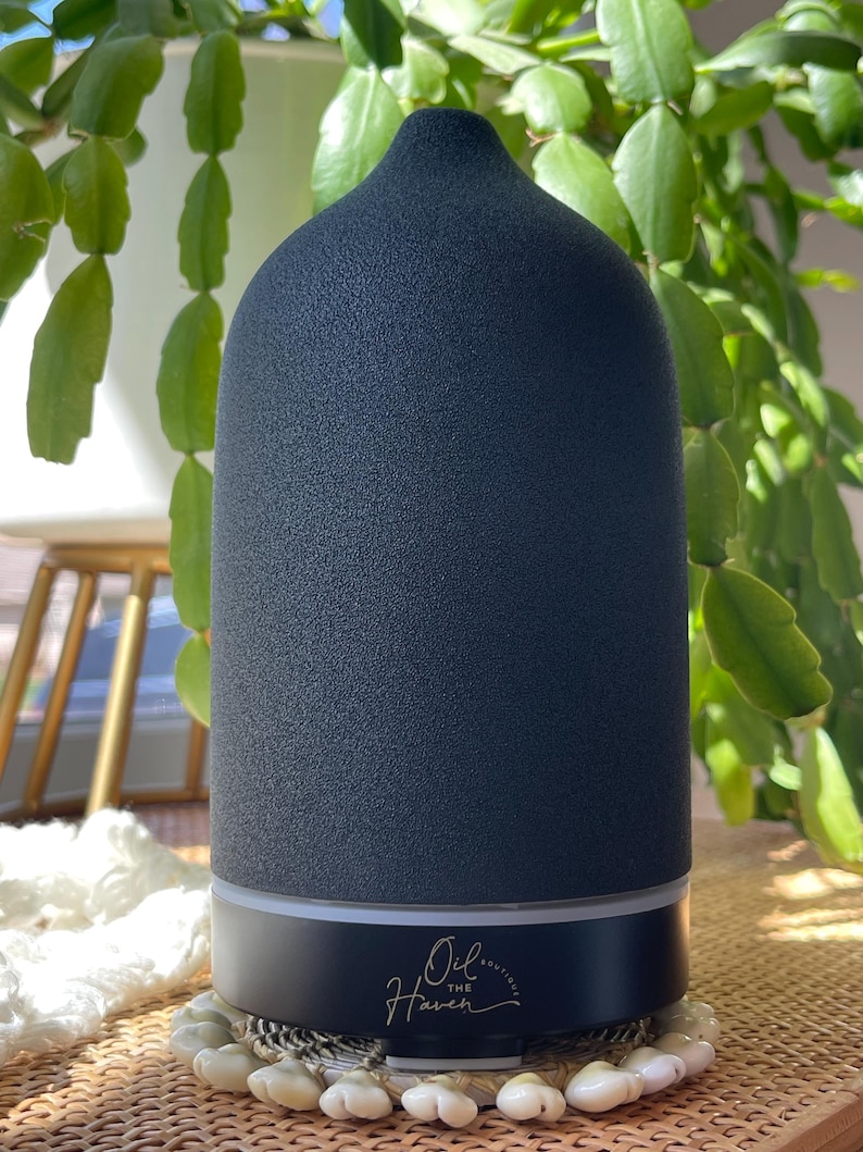 Puede incluir: Un difusor de aceites esenciales gris oscuro con una superficie texturizada y una parte superior redondeada. La base es negra con el texto "Oil the Haven" en escritura dorada. El difusor se encuentra sobre una superficie de cuentas blancas, con plantas verdes en el fondo.