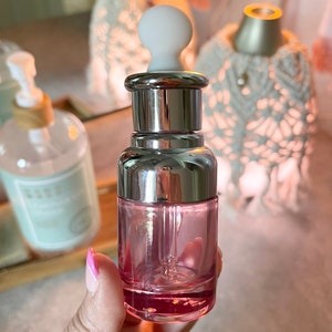 Puede incluir: Una botella de vidrio transparente con una tapa plateada y un gotero blanco. La botella est&aacute; llena de un l&iacute;quido rosa.