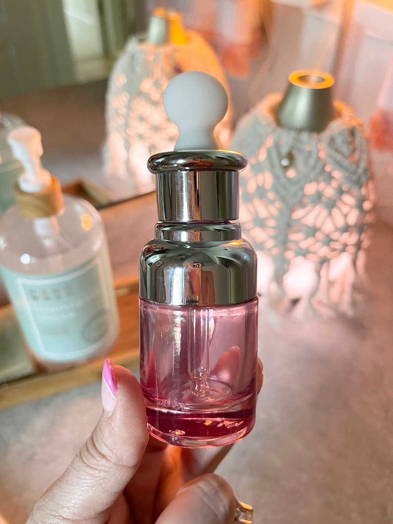 Puede incluir: Una botella de vidrio transparente con una tapa plateada y un gotero blanco. La botella est&aacute; llena de un l&iacute;quido rosa.