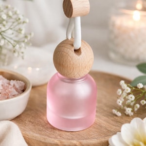 May include: A small, round, frosted pink glass bottle with a light-colored wooden cap. The bottle is suspended by a white cord that features a small, cylindrical wooden bead for adjustment, designed for hanging as a car air freshener.